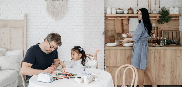 Father and daughter bond over crafts while mother prepares food in a cozy home setting.