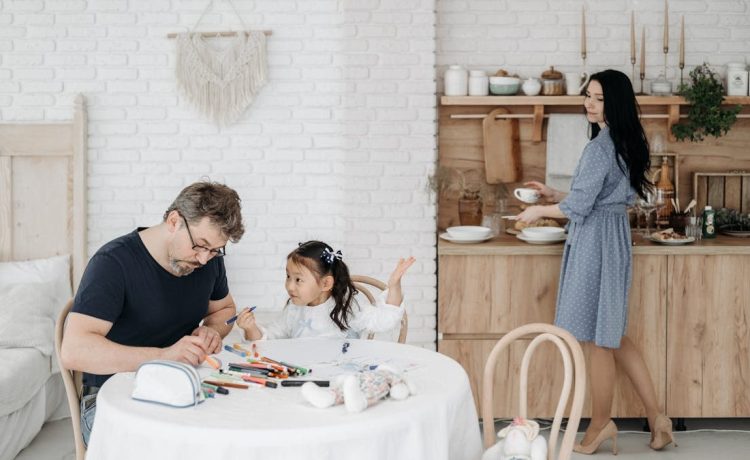 Father and daughter bond over crafts while mother prepares food in a cozy home setting.